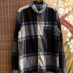 May include: A long-sleeved plaid shirt in black, gray, and beige, with teal and red accent stripes. The shirt has a button-down front, a chest pocket, and a collared neckline. The shirt is hanging on a hook.