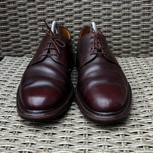 May include: A pair of burgundy leather dress shoes with a classic Oxford design. The shoes have a rounded toe, a lace-up closure, and a dark brown sole. The shoes are displayed on a woven wicker surface.