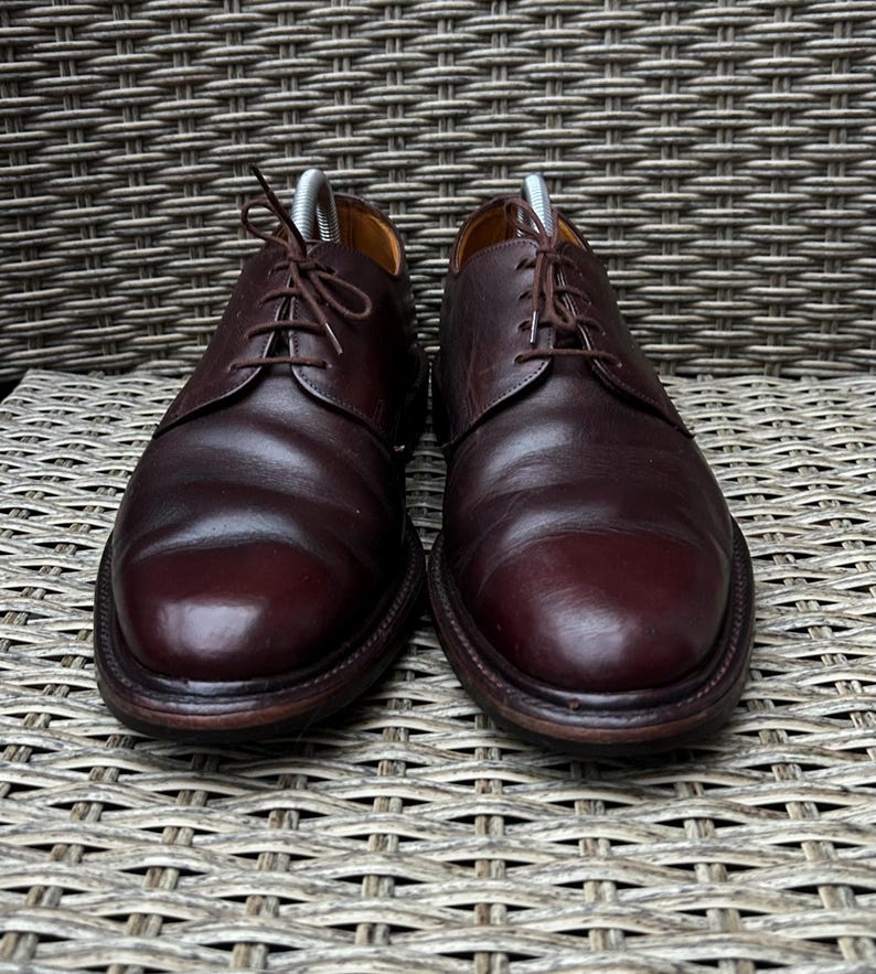May include: A pair of burgundy leather dress shoes with a classic Oxford design. The shoes have a rounded toe, a lace-up closure, and a dark brown sole. The shoes are displayed on a woven wicker surface.