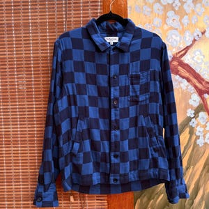 Pode incluir: Casaco xadrez azul marinho e azul, com fecho de botões, bolso no peito e mangas compridas. O casaco está pendurado num cabide preto. A etiqueta na gola diz "You Must Create".