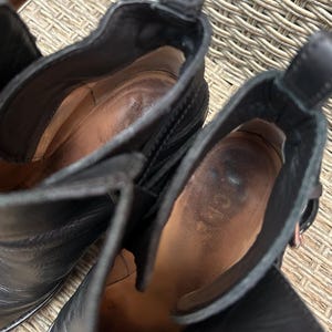 May include: Close-up of two black leather ankle boots. The interior of the boots is visible, showing a worn, brown leather insole. The boots have a pull tab at the back and a buckle detail. The brand name "ACNE" is visible inside one boot.