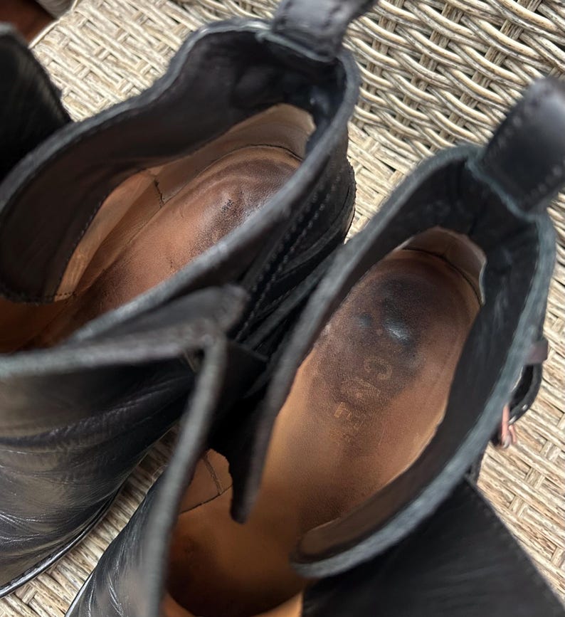 May include: Close-up of two black leather ankle boots. The interior of the boots is visible, showing a worn, brown leather insole. The boots have a pull tab at the back and a buckle detail. The brand name "ACNE" is visible inside one boot.