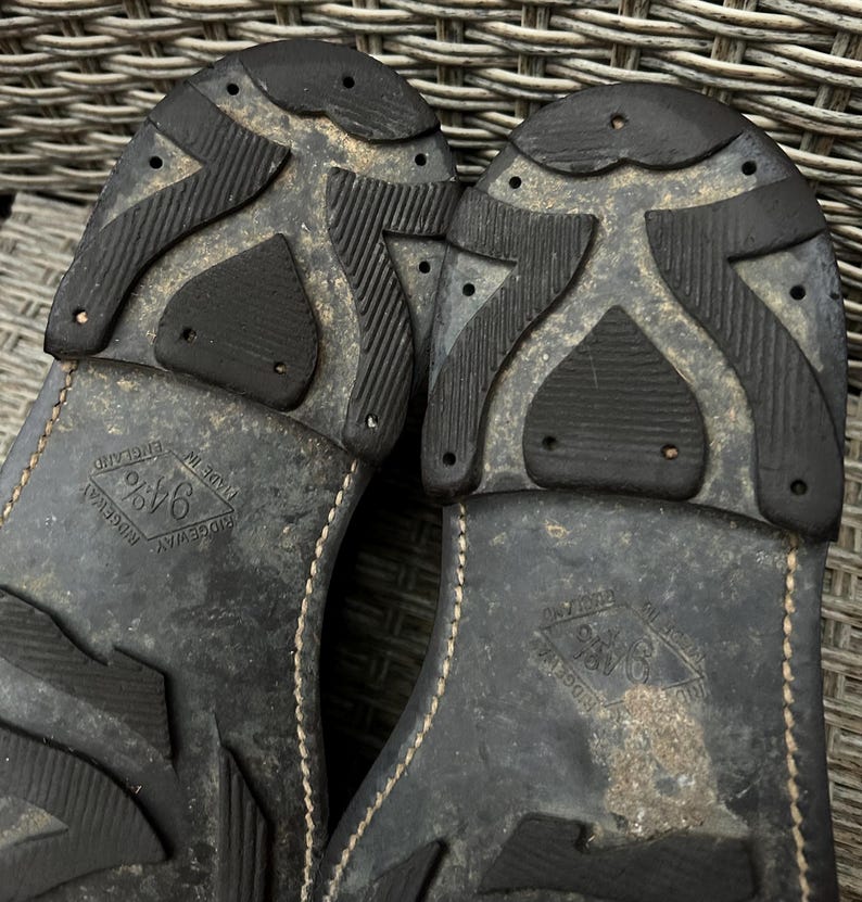 May include: Close-up of the soles of two black leather shoes. The soles have a textured, patterned design with the words "Ridgeway" and "Made in England" visible. The shoes appear worn.