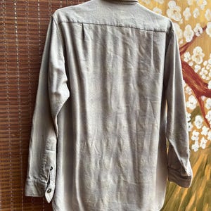 以下が含まれることがあります： フックに掛けられた長袖の薄茶色のシャツ。襟、ボタン付きの袖口、カーブした裾が特徴です。生地は柔らかく織られた素材のようです。背景には竹製のブラインドと絵画があります。