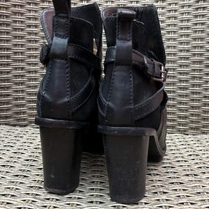May include: A pair of black ankle boots with a stacked heel. The boots feature a buckle and strap detail around the ankle and a pull tab at the back. The heels measure approximately 3 inches.