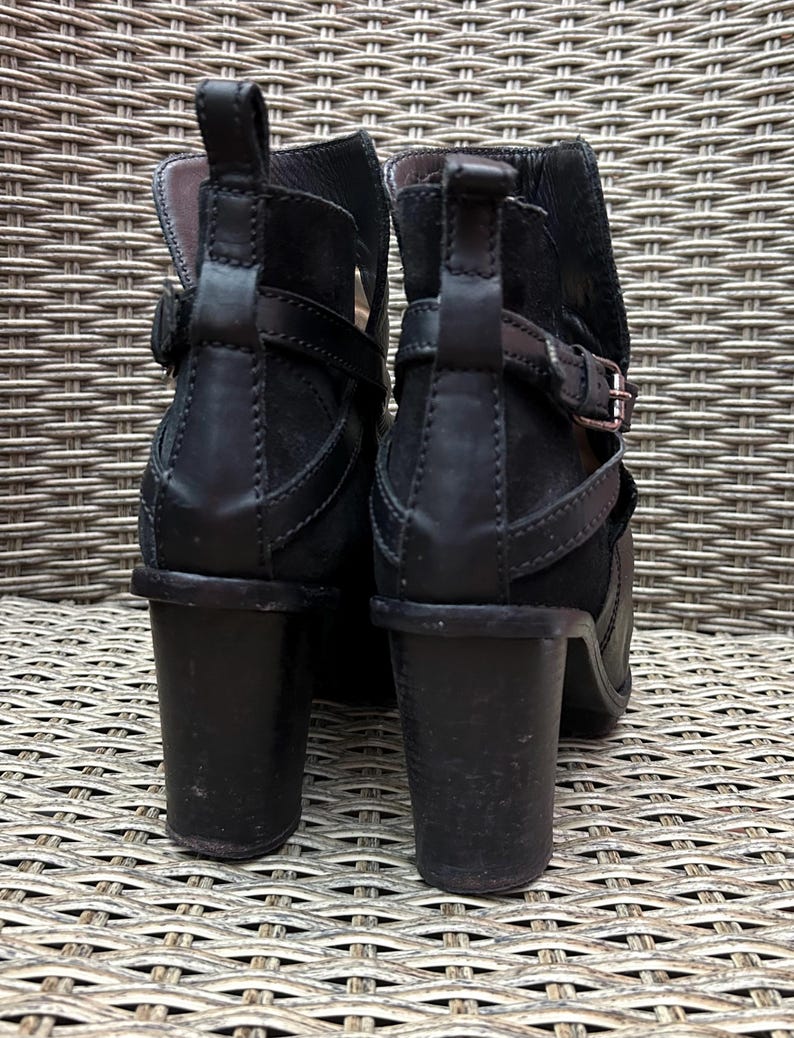 May include: A pair of black ankle boots with a stacked heel. The boots feature a buckle and strap detail around the ankle and a pull tab at the back. The heels measure approximately 3 inches.
