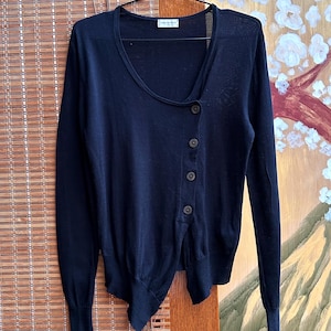 May include: A dark navy blue cardigan with an unusual asymmetrical button fastening. The long-sleeved sweater has a scoop neckline and is hanging on a black hanger. The cardigan is made of a soft, lightweight material.