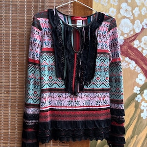 Op de afbeelding: Een top met lange mouwen met een kleurrijk geometrisch patroon in rood, zwart, roze en teal. De top heeft een sleutelgat-halslijn met zwarte kanten afwerking en kanten details aan de manchetten en zoom. Het kledingstuk hangt aan een witte hanger.