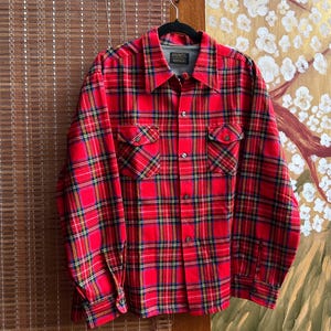 May include: A long-sleeve button-up shirt featuring a vibrant red plaid pattern with thin yellow and blue accent lines. The shirt includes a pointed collar, two chest pockets with buttoned flaps, and a full-length front button closure.