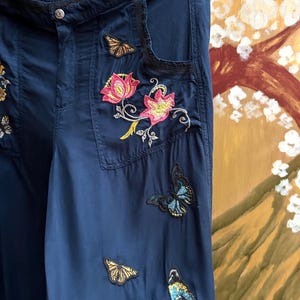 May include: Navy blue trousers adorned with embroidered floral and butterfly motifs. The trousers have a button fastening and pockets. The embroidery incorporates pink, yellow, and blue, enhancing the garment's decorative appeal.