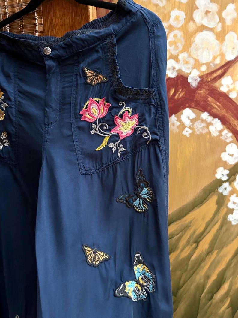 May include: Navy blue trousers adorned with embroidered floral and butterfly motifs. The trousers have a button fastening and pockets. The embroidery incorporates pink, yellow, and blue, enhancing the garment's decorative appeal.