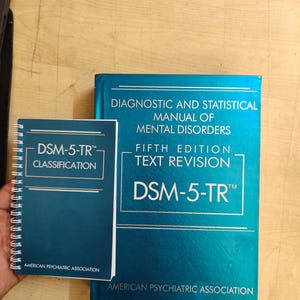 May include: Two blue books, one spiral-bound, and one larger, titled "DSM-5-TR Classification" and "Diagnostic and Statistical Manual of Mental Disorders, Fifth Edition, Text Revision." Both books are from the American Psychiatric Association.