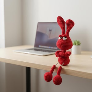 May include: A handmade red crochet bunny doll with crossed arms and long legs with red ball feet. The bunny has white eyes and is sitting on a wooden desk. A laptop and small potted plant are in the background.