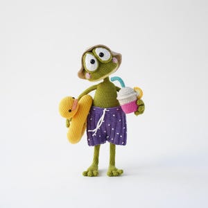 May include: A crocheted frog wearing a hat, purple polka-dot swim trunks, and holding a yellow pool float and a drink. The frog is green with large white-eyed features and a tan hat. The drink is pink, white, and blue.