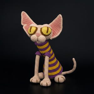 Könnte beinhalten: Handgemachte Häkelkatzenpuppe mit großen gelben Augen und einem gestreiften lila und gelben Hemd. Die Katze hat lange Beine und große Ohren. Die Katze sitzt auf einer schwarzen Oberfläche.