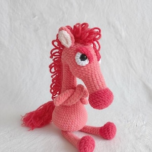 Könnte beinhalten: Ein handgefertigtes, rosa gehäkeltes Pferd mit roter Mähne und Schweif. Das Pferd hat einen langen Hals, einen runden Körper und kleine Beine. Die Augen sind weiß mit schwarzen Pupillen. Das Pferd sitzt auf einer weißen Oberfläche.