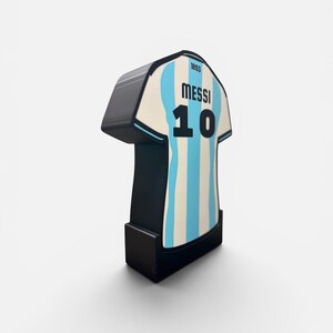 May include: A three-dimensional, upright display piece shaped like a soccer jersey with light blue and white vertical stripes. The front features the text "MESSI" above the number "10" and the small number "1893" near the collar, all in black.
