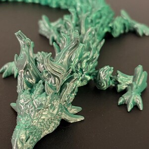 May include: A detailed, articulated green dragon figurine. The dragon has a textured, scaly appearance with intricate details on its head, body, and limbs. The dragon is lying down on a dark surface. The dragon's body is approximately 12 inches long.