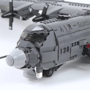 May include: A grey model aeroplane made from interlocking plastic bricks. The model features four propellers, the text "AIR-GUNSHIP" and the number "130" on the fuselage. The plane has a detailed cockpit and a white nose cone.
