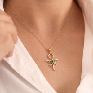 May include: A gold-colored pendant necklace featuring an Ankh symbol with a green gemstone at its center. The necklace is worn against a white shirt, showcasing the pendant's design and color.