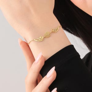 Könnte beinhalten: Ein filigranes goldenes Armband mit einem Blumendesign und zwei Flügelpaaren. Das Armband wird am Handgelenk getragen und zeigt seine filigranen Details und sein elegantes Design. Ein schönes Schmuckstück.