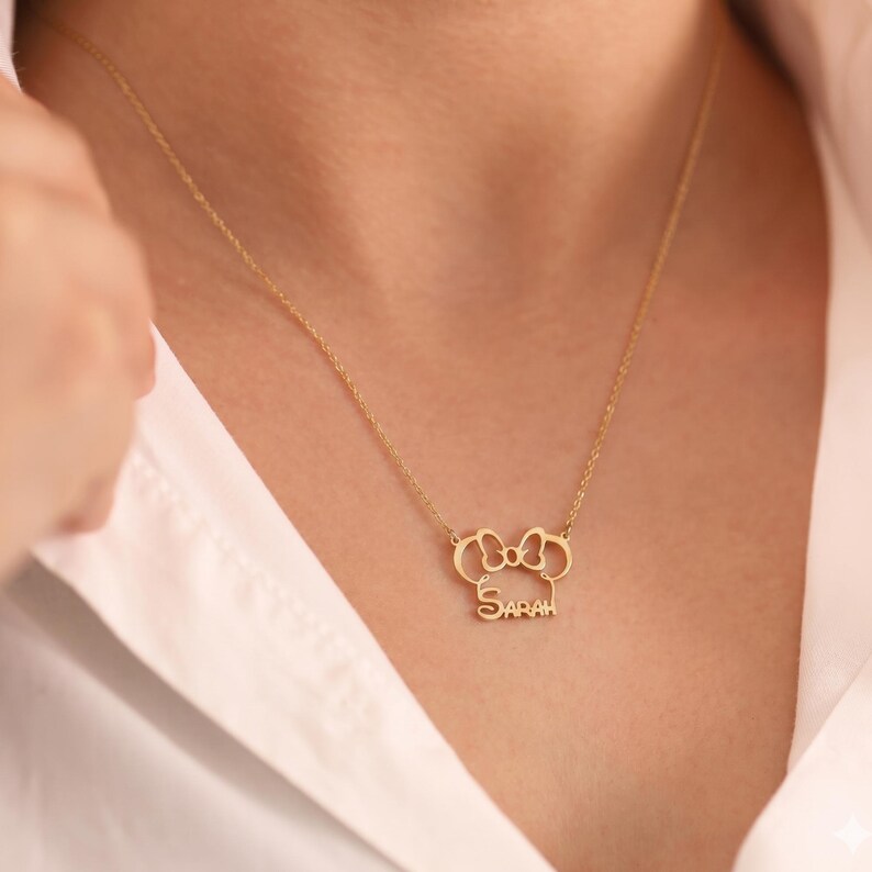 May include: A gold-colored necklace with a pendant shaped like a bow and two circles, with the name "SARAH" below. The necklace is worn on a person wearing a white shirt.