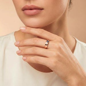 Puede incluir: Anillo de oro rosa con una piedra azul y pequeñas piedras blancas. El anillo está en un dedo, mostrando su diseño y color. El anillo es una pieza de joyería.