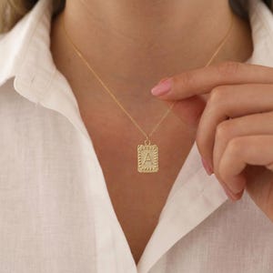 May include: A gold-colored necklace with a rectangular pendant featuring the letter "A". The pendant has a textured border and a delicate chain. The necklace is worn against a white shirt.