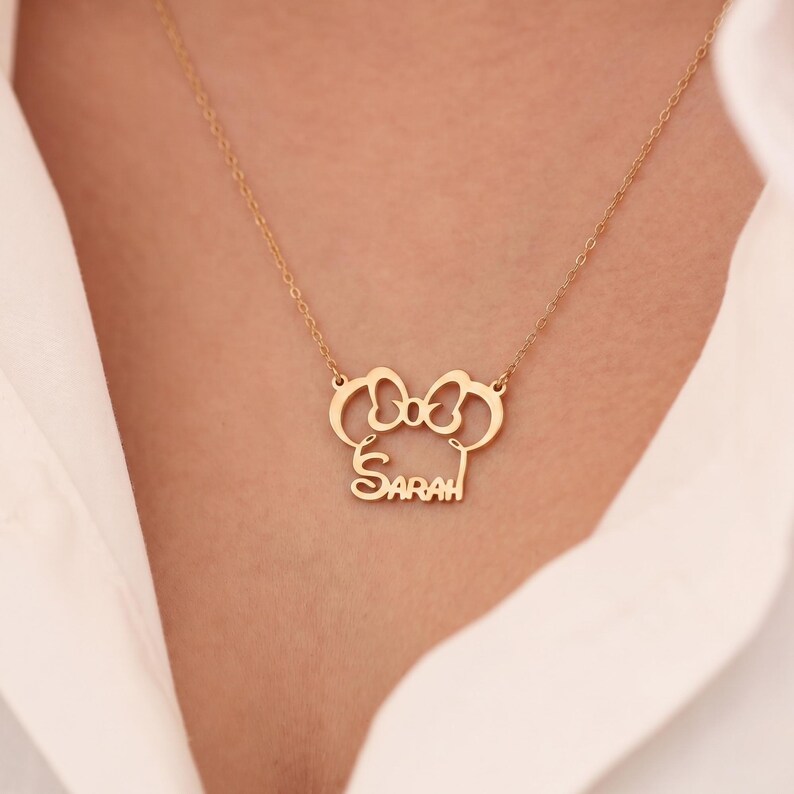 May include: A gold-colored necklace with a bow-shaped pendant and the name "Sarah" in a script font. The necklace is worn against a white shirt. The pendant is approximately 1 inch wide.