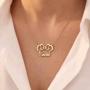 May include: A gold-colored necklace with a pendant featuring a bow design and the name "Sarah" in a delicate font. The necklace is worn against a white shirt, highlighting the pendant's details.