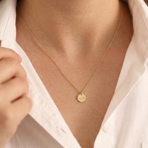 May include: A gold-colored necklace with a delicate chain and a small, round pendant. The pendant is inscribed with the name "nick" in a delicate script. The necklace is worn against a white shirt.