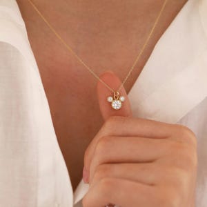 May include: A delicate gold necklace featuring a pendant with a central round, clear gemstone surrounded by smaller gemstones, creating a Mickey Mouse-inspired design. The necklace is displayed against a white shirt.