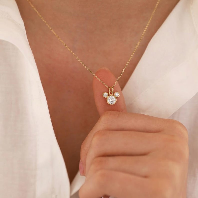 May include: A delicate gold necklace featuring a pendant with a central round, clear gemstone surrounded by smaller gemstones, creating a Mickey Mouse-inspired design. The necklace is displayed against a white shirt.