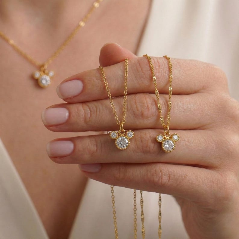 May include: Gold-toned necklaces featuring a Mickey Mouse-inspired pendant with a large, round, clear gemstone center and smaller side stones. The necklaces are displayed against a neutral background, showcasing the jewelry's delicate design and sparkle.