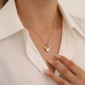 May include: A gold-colored necklace with a pendant featuring a three-circle design, with a large, clear, round stone in the center and two smaller stones on either side. The necklace is worn against a white shirt.