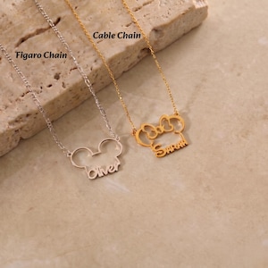 May include: Two necklaces with pendant designs. One is silver with a Mickey Mouse head shape and the name "Oliver". The other is gold with a bow shape and the name "Sarah". Both have chain styles labeled "Figaro Chain" and "Cable Chain".