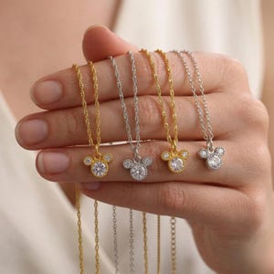 May include: Four delicate necklaces with Mickey Mouse-shaped pendants. Two are gold-toned, and two are silver-toned. Each pendant features a central clear gemstone surrounded by smaller stones. The chains are thin and twisted.