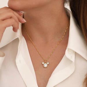 May include: A gold-colored necklace with a pendant shaped like a mouse head. The pendant features a large, round, clear gemstone surrounded by smaller gemstones. The necklace is worn against a white collared shirt.