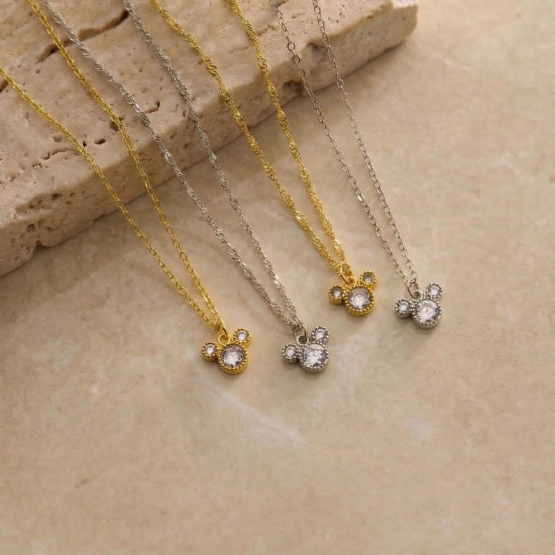 May include: Several delicate necklaces with a Mickey Mouse-shaped pendant. The pendants are adorned with clear, sparkling stones. The chains are either gold or silver-toned, and the background is a neutral beige.
