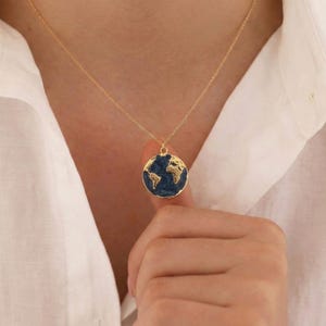 May include: A gold-colored necklace featuring a pendant shaped like a globe. The globe pendant has a dark blue background with gold-colored continents. The necklace is worn against a white shirt.