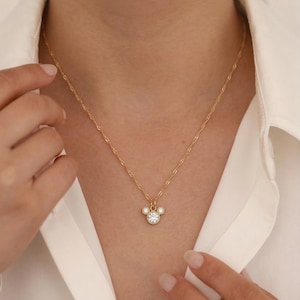 May include: Gold-colored necklace with a pendant featuring a three-circle design, with a large central clear stone and smaller stones around the edges. The necklace is worn against a white shirt.