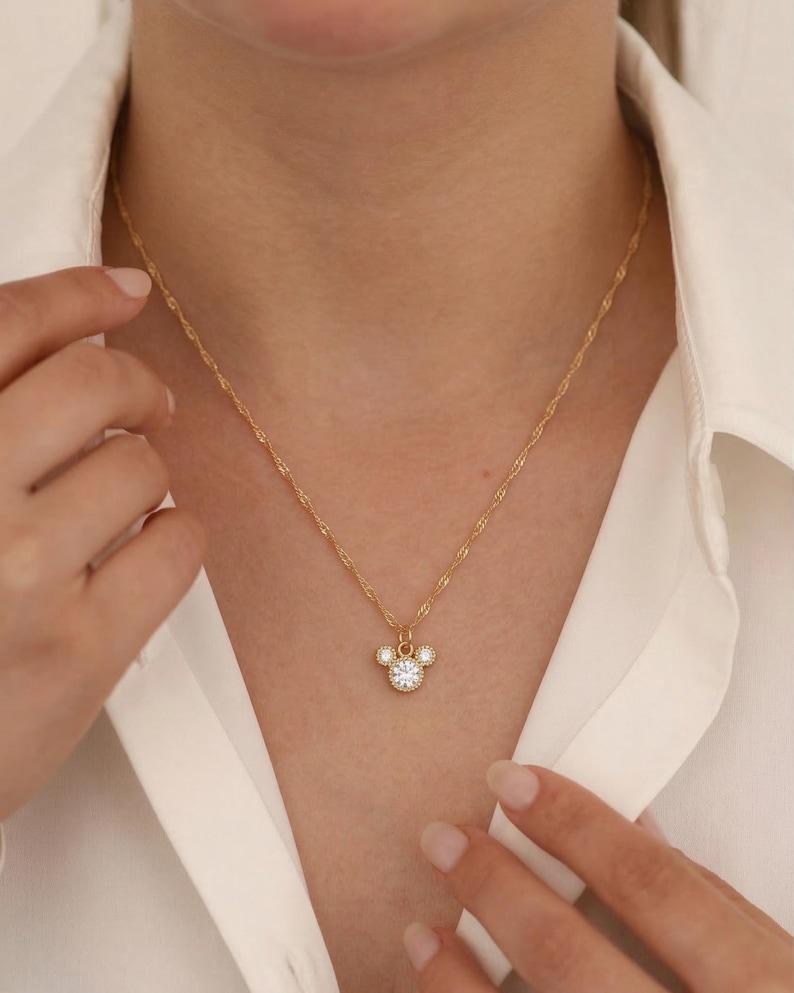 May include: Gold-colored necklace with a pendant featuring a three-circle design, with a large central clear stone and smaller stones around the edges. The necklace is worn against a white shirt.