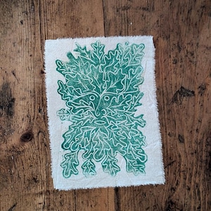 May include: A square piece of off-white fabric featuring a green Green Man print. The design depicts a face within a leafy pattern. The fabric has a raw edge, set against a wooden background.