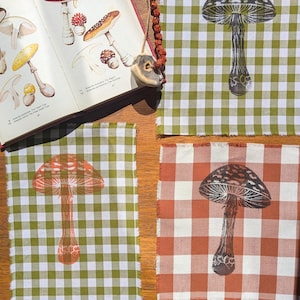 May include: Four fabric squares featuring mushroom designs on a wooden surface. Two squares have green and white gingham, one with red and white gingham. An open book with mushroom illustrations is also present.