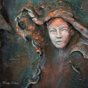 May include: A close-up of a textured, three-dimensional sculpture of a woman's face. The sculpture is painted in shades of green, brown, and copper. The woman's face is partially obscured by a flowing piece of fabric. The sculpture is signed "Art by Dr Franky Dolan".
