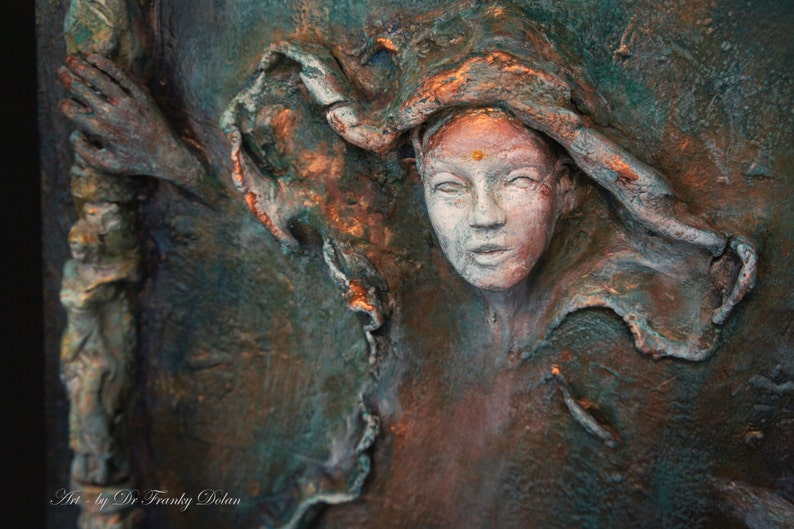 May include: A close-up of a textured, three-dimensional sculpture of a woman's face. The sculpture is painted in shades of green, brown, and copper. The woman's face is partially obscured by a flowing piece of fabric. The sculpture is signed "Art by Dr Franky Dolan".