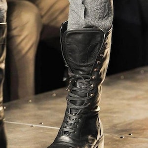 May include: Black leather knee-high boots with a lace-up design and small heel. The boots have a classic, rugged style, with multiple eyelets and a sturdy sole. The boots are worn with grey trousers.
