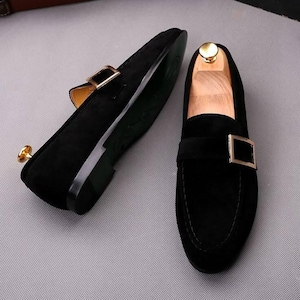 K&ouml;nnte beinhalten: Ein Paar schwarze Wildleder-Loafer mit goldfarbenen Metallschnallen. Die Schuhe haben ein klassisches Design mit runder Spitze und niedrigem Absatz. Die Sohlen sind dunkelgr&uuml;n. Die Schuhe werden auf einer hellgrauen Oberfl&auml;che pr&auml;sentiert.