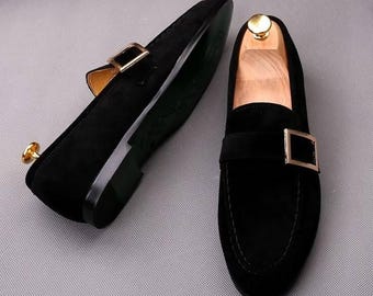 May include: A pair of black suede loafers with gold-colored metal buckles. The shoes have a classic design with a rounded toe and a low heel. The soles are a dark green color. The shoes are displayed on a light gray surface.