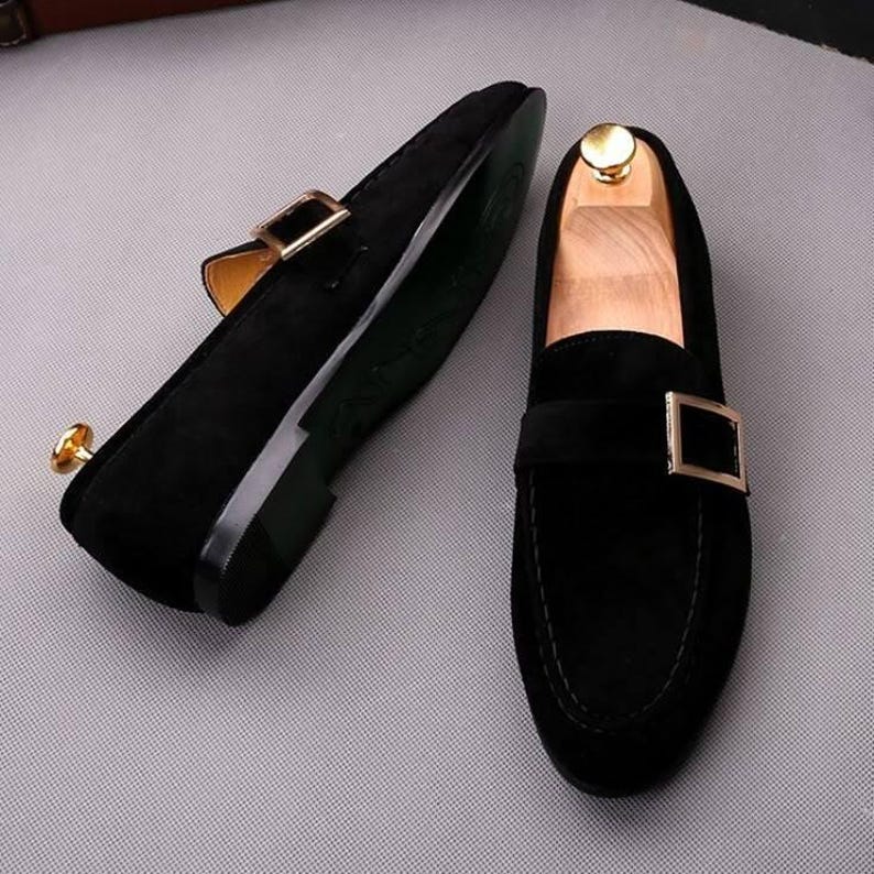 K&ouml;nnte beinhalten: Ein Paar schwarze Wildleder-Loafer mit goldfarbenen Metallschnallen. Die Schuhe haben ein klassisches Design mit runder Spitze und niedrigem Absatz. Die Sohlen sind dunkelgr&uuml;n. Die Schuhe werden auf einer hellgrauen Oberfl&auml;che pr&auml;sentiert.